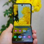 A hand holding a foldable smartphone reflecting yellow flowers on screen.